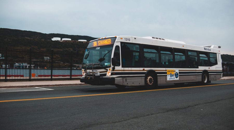 Big gray bus driving on asphalt highway along metal fence near water against cloudless sky with mountains in distance outside