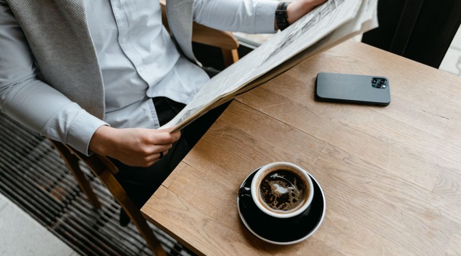 An adult male enjoying his coffee while reading a newspaper at a cafe with his smartphone nearby. — Generate More Engagement