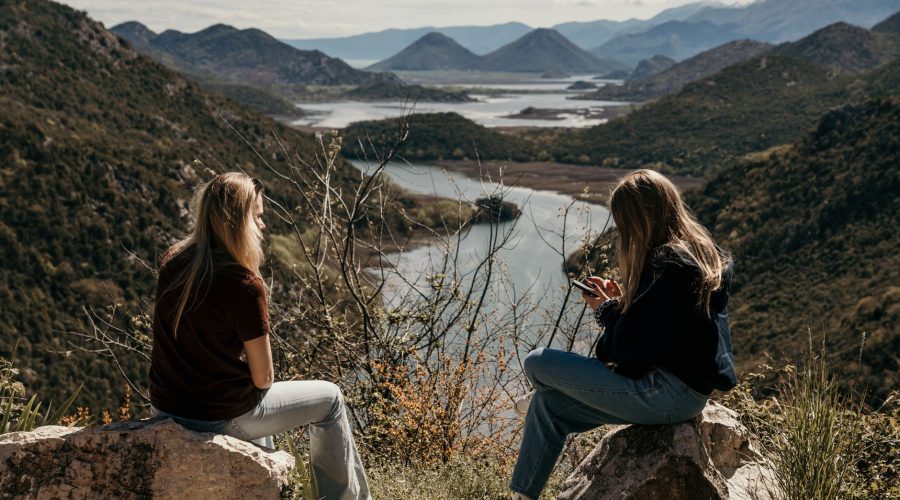 Two women sitting on rocks, overlooking a beautiful mountain and river landscape. — Hotels & Timeshares Versus. Holiday Renta