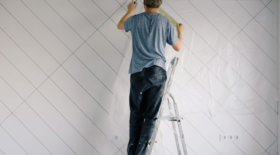Full body back view of faceless male in shirt and dirty pants repainting wall while standing on ladder in bright empty room i